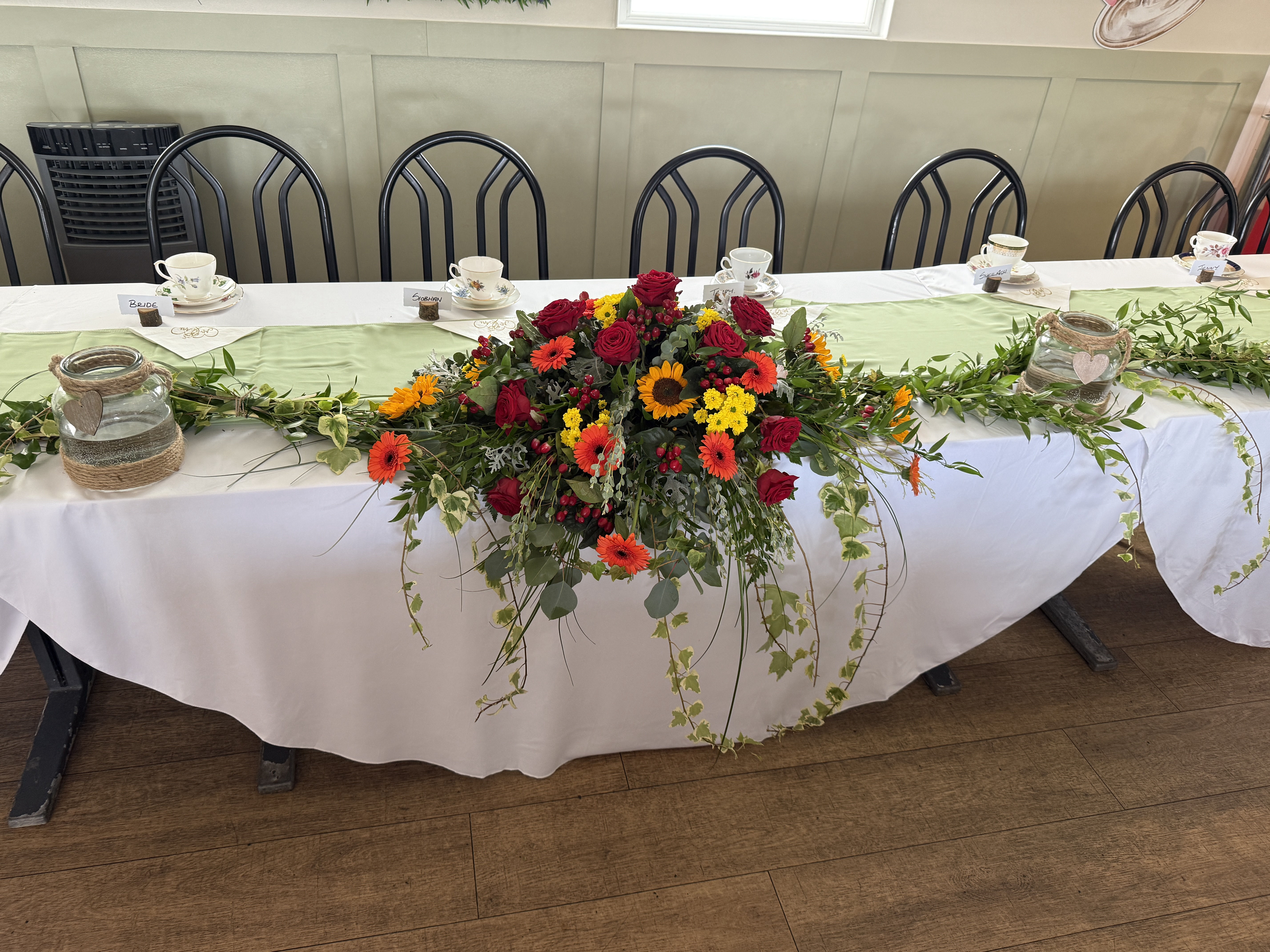 A long, low arrangement sitting at the front of a long table created in shades of yellow, orange and deep reds, including Sunflowers and Red Roses, with ivy trailing.