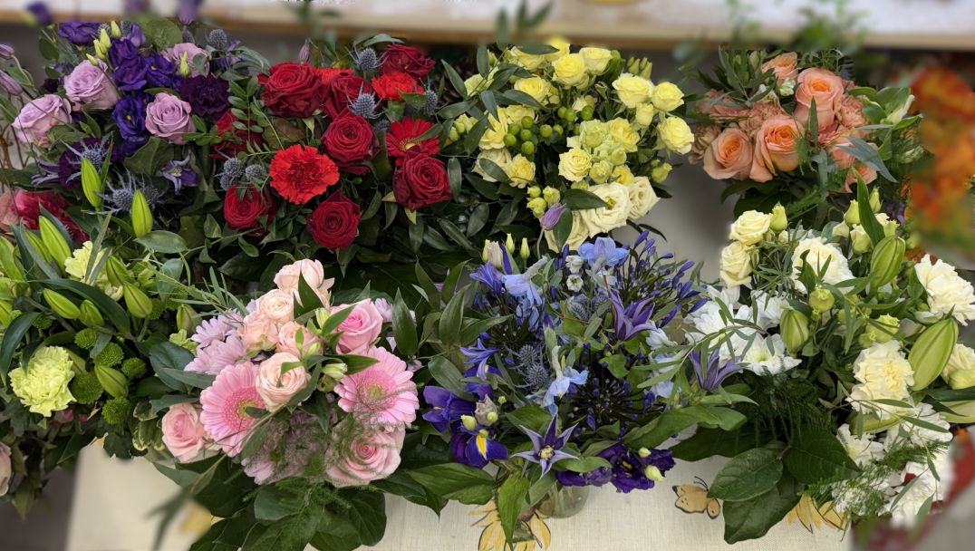 MobiriseA photo showing eight jam jars filled with flowers, with a row of four behind another row of four. Each jar has a different colour. There is a red, purple, all green, lemon, pink, cream, peach and blue one. The flowers used include roses, eryngium, eustoma, lilies, carnations, clematis and other seasonal blooms. 
